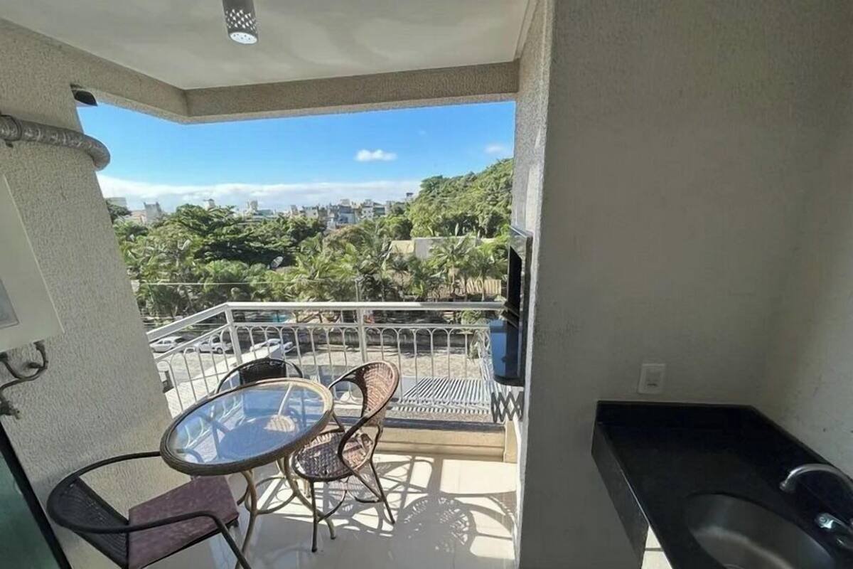 A balcony with a glass table and chairs, a sink, and a view of a cityscape with greenery.