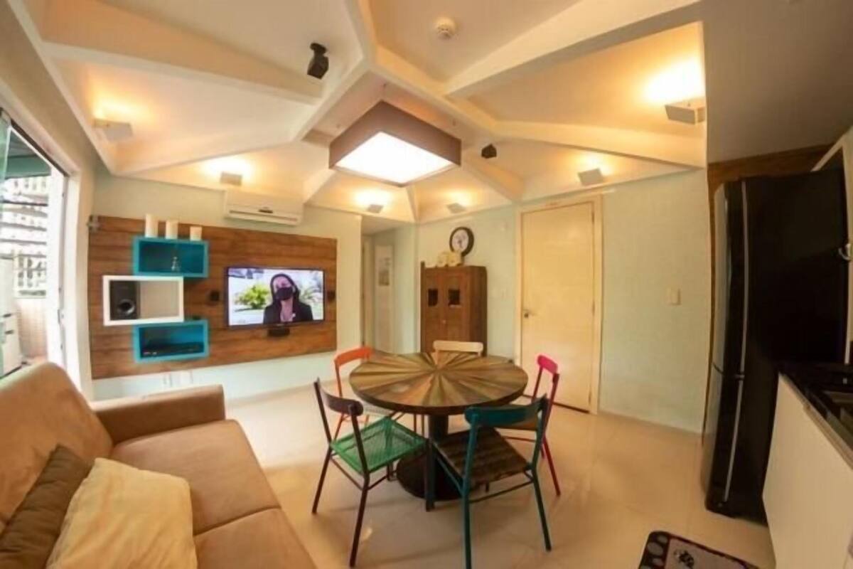 A modern living room with a round dining table, colorful chairs, a sofa, and a flat-screen TV mounted on the wall.