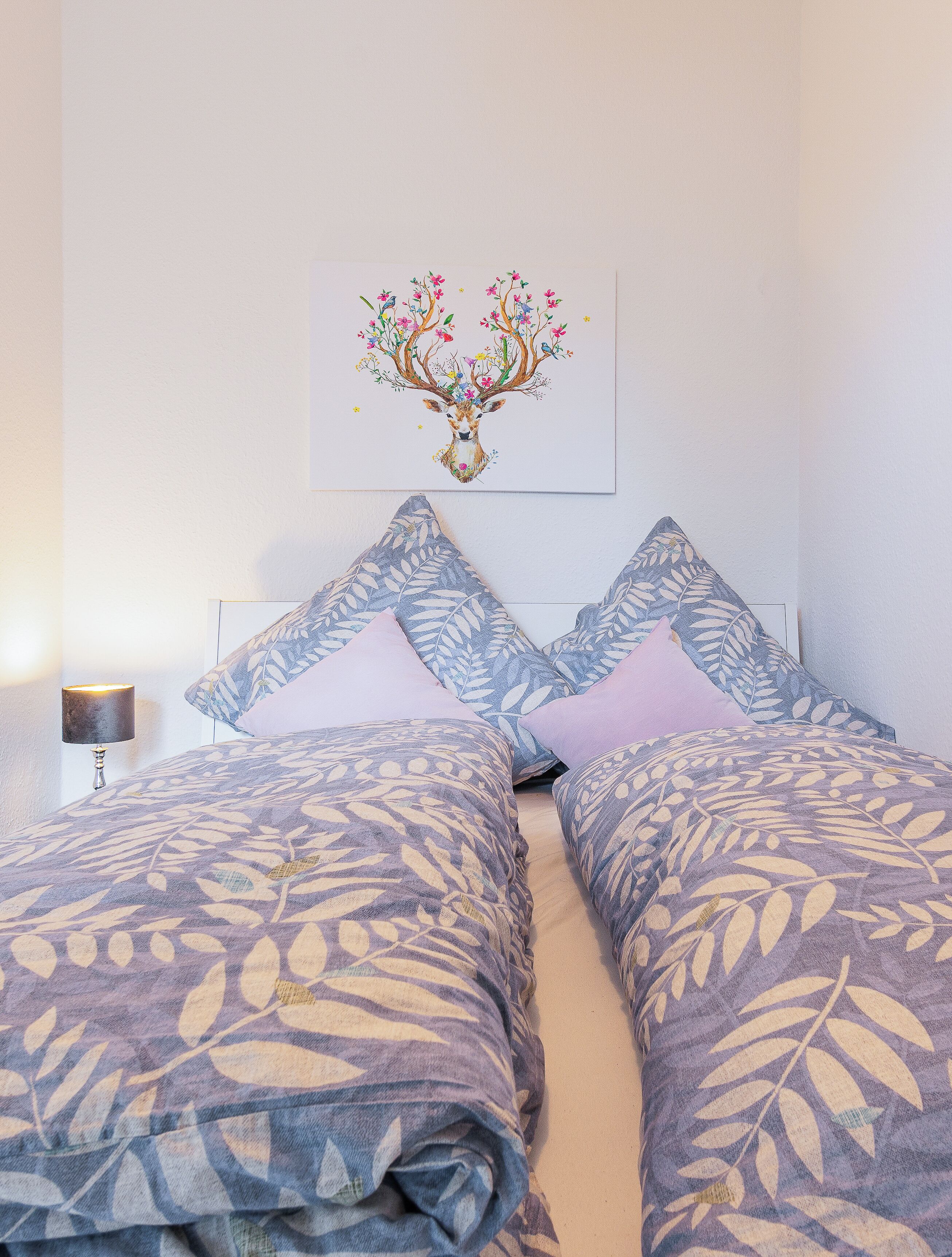 Un lit bien fait, avec une literie à motifs, une lampe fixée au mur et une œuvre d’art encadrée représentant un cerf entouré de fleurs.