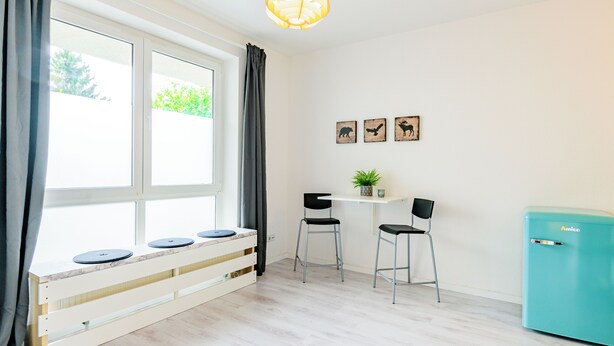 A room with a window, a small table with chairs, a plant, and a refrigerator. - Modern apartment with parking space & excellent connections - ideal for two (Magdeburg)