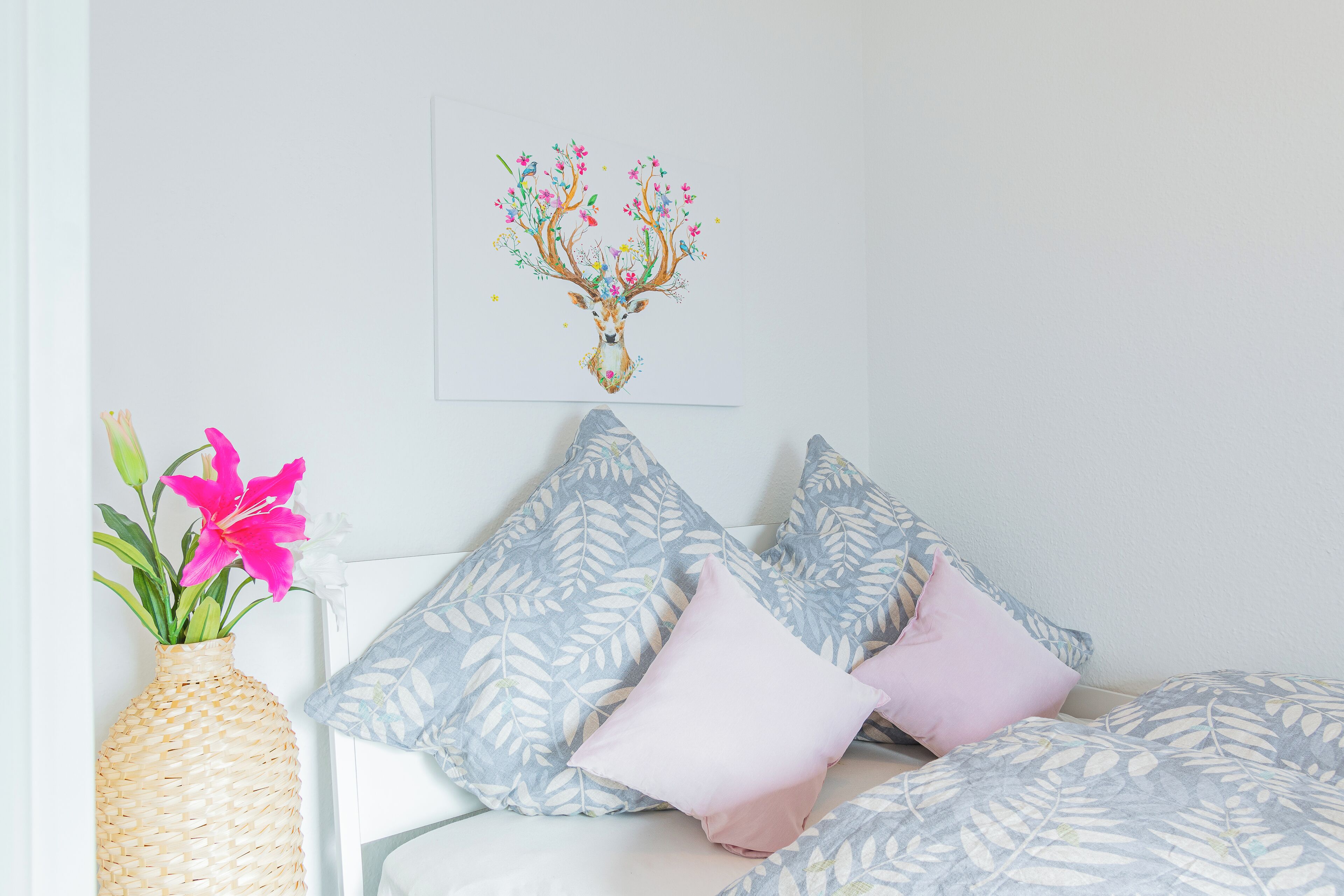 Un dormitorio con cama, almohadas, un cuadro de un ciervo y un florero con flores.