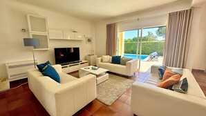 Een moderne woonkamer met witte meubels, een flatscreen televisie en uitzicht op een zwembad.