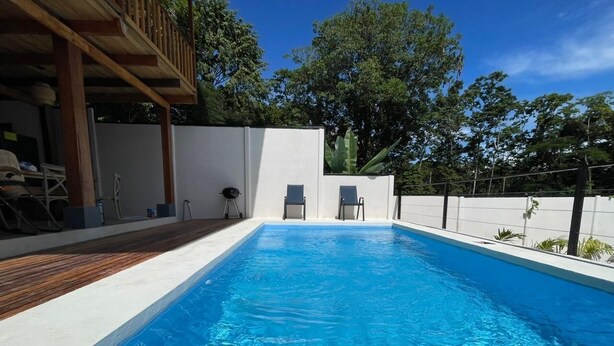 Une piscine rectangulaire avec une zone de terrasse en bois d’un côté, entourée d’un mur blanc et bordée d’arbres en arrière-plan.