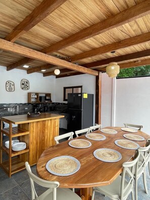 A wooden dining table with woven placemats, surrounded by white chairs, in a kitchen area with a wooden ceiling and a black refrigerator.