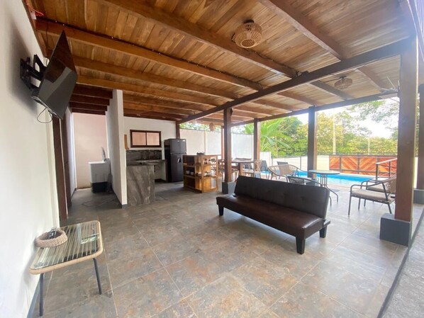 A spacious living area with a wooden ceiling, a sofa, a TV, a kitchen area, and a pool view.