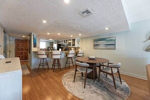 A dining area with a round table and bar stools, a kitchen in the background, and a door leading outside. - Vitamin Sea Condo (Jacksonville Beach)