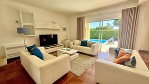 Een moderne woonkamer met witte meubels, een flatscreen televisie en uitzicht op een zwembad.