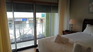 Une chambre d’hôtel avec un grand lit, une table de chevet avec une lampe et une vue sur la ville grâce à de grandes fenêtres du sol au plafond.