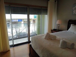 A hotel room with a large bed, a nightstand with a lamp, and a view of the city through floor-to-ceiling windows. - Spacious and comfortable apartment Ponta Delgada (Ponta Delgada)