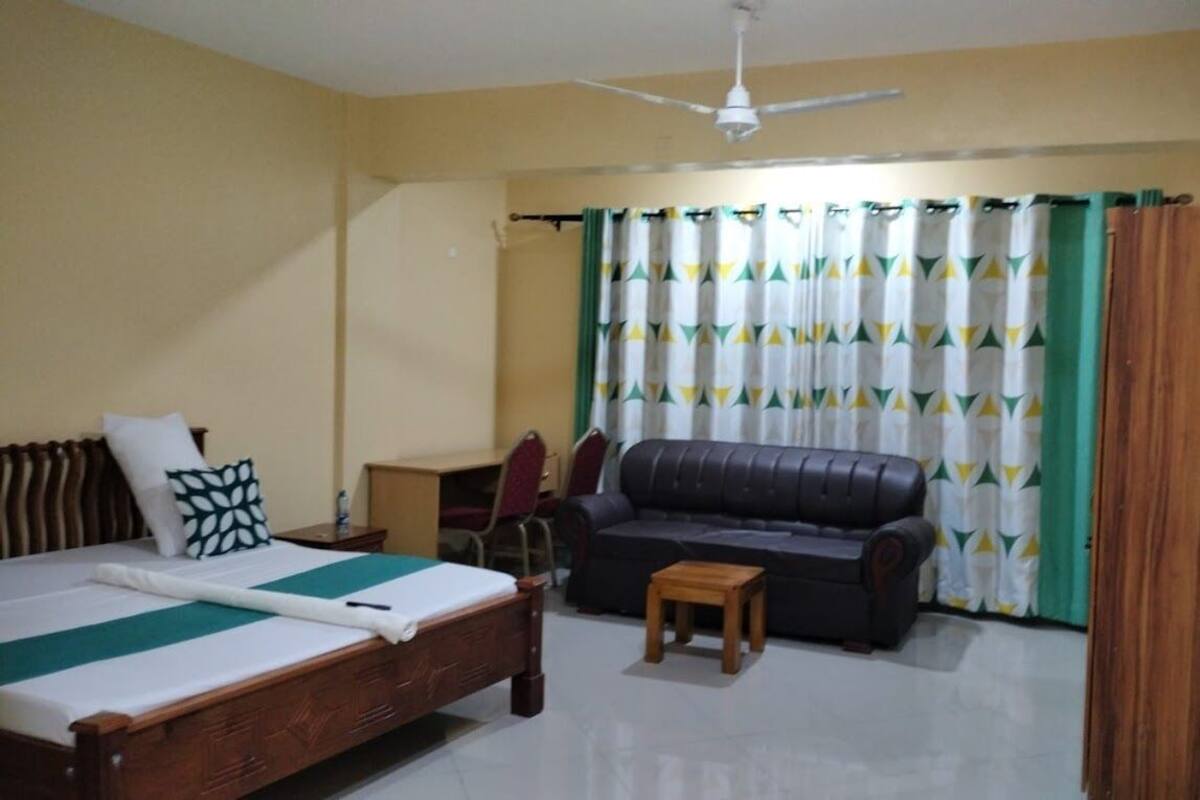 A bedroom with a bed, a sofa, a small table, a desk, and a television on the wall.