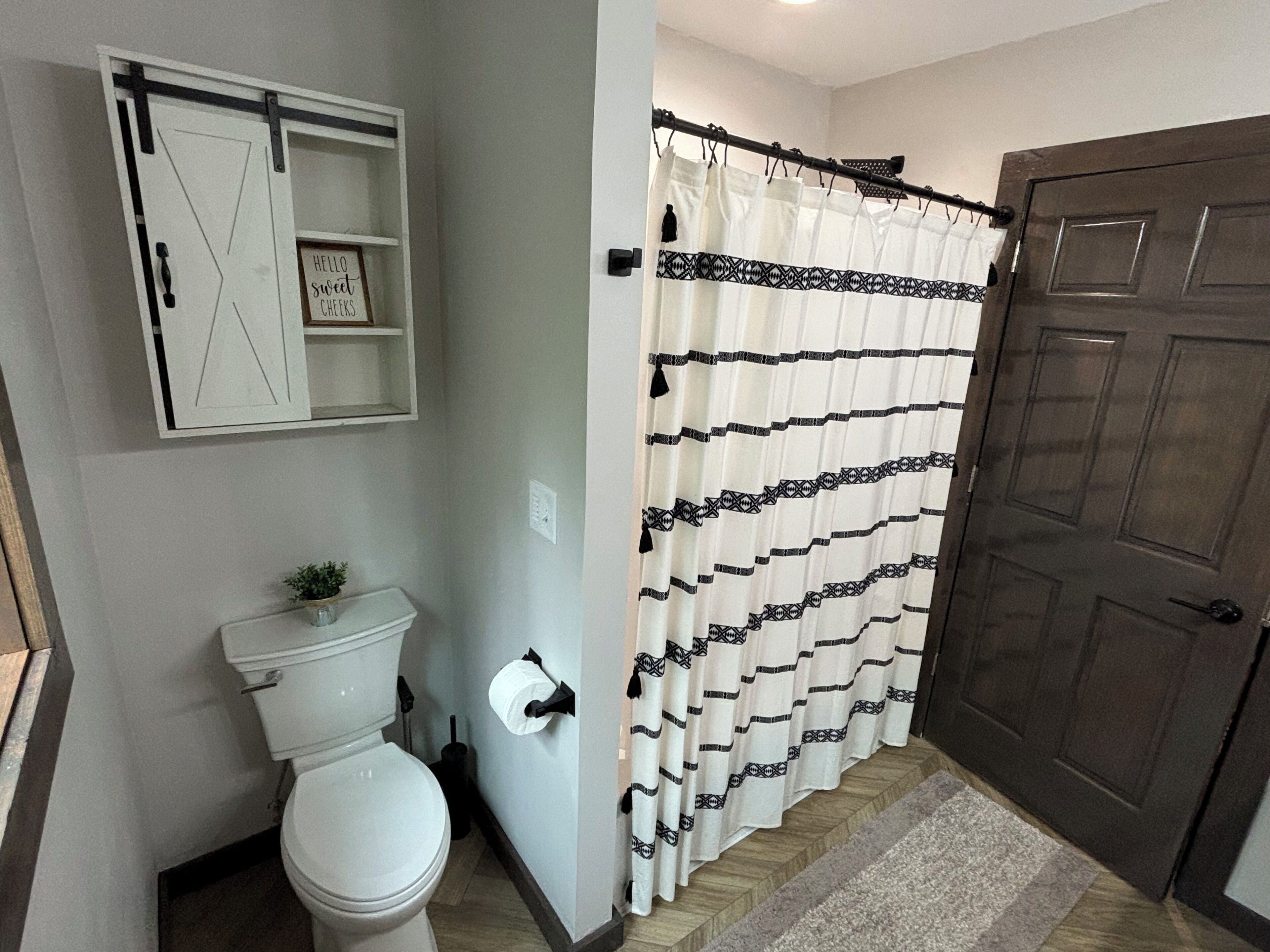 A bathroom with a toilet, a shower curtain, a door, and a shelf with a sign.