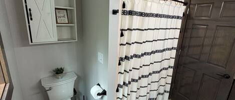 A bathroom with a toilet, a shower curtain, a door, and a shelf with a sign.