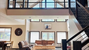A spacious living area with a high ceiling, a large clock, and a staircase leading to an upper level.