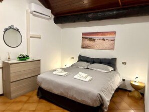 Une chambre à coucher comprenant un lit, une commode, un climatiseur, un miroir fixé au mur, un tableau représentant une plage et une lampe de chevet.