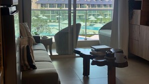 Ett hotellrum med ett stort fönster med utsikt över en pool och en byggnad med flera tak. Det finns en soffa, ett litet bord och en stol.
