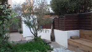 Un pati amb una banca de fusta, un arbre petit i una paret blanca.