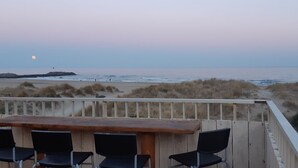 Una terrassa de fusta amb una taula i cadires, amb vista a una platja de dunes i un far a la distància.