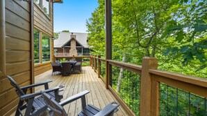 Una terraza de madera con mobiliario exterior, una chimenea de piedra y vistas a una casa rodeada de árboles.