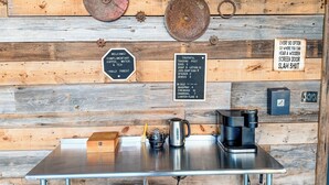 Pared de madera rústica con carteles que indican café, agua y té de cortesía, una máquina de café y un hervidor.