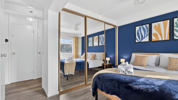 Ein Schlafzimmer mit einem großen Spiegel, einem Bett mit blauer Bettdecke, einem Nachttisch mit Lampe und Kunstwerken an der Wand.