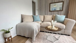 A living room with a beige sectional sofa, a small wooden coffee table, a white vase with dried flowers, and a potted plant. - Cornish coastal escape, close to the beach, shops, cafes and restaurants. (Bude)