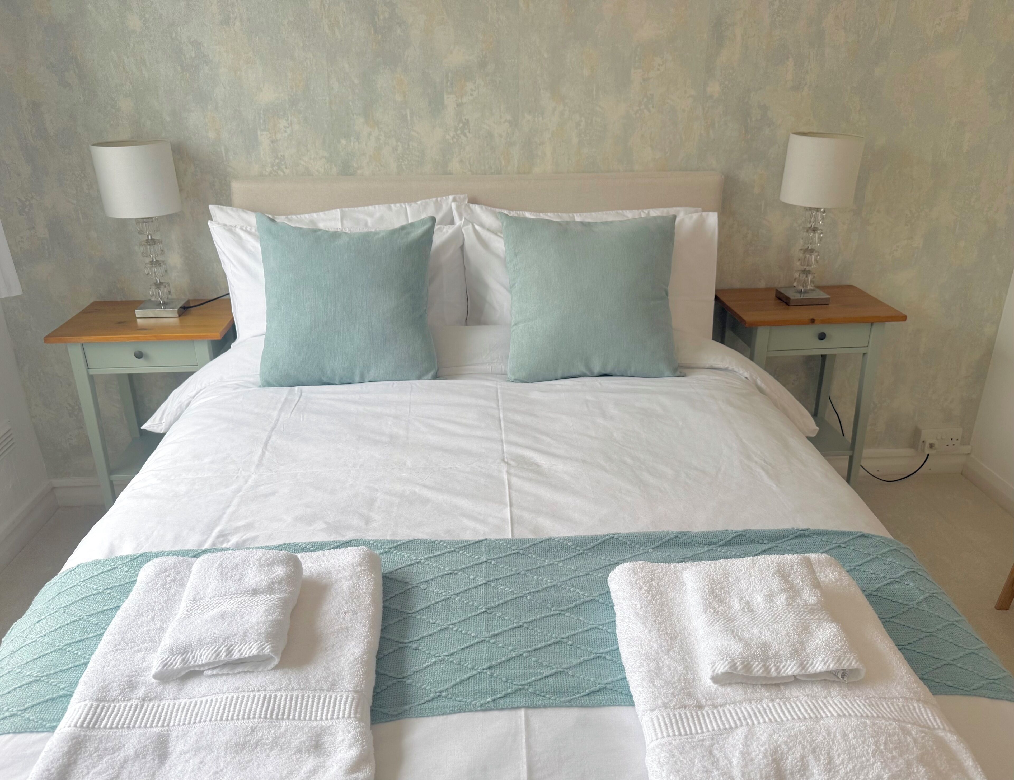 A neatly made bed with a white and teal bedspread, two matching pillows, and two bedside tables with lamps and books.