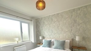 A bedroom with a large bed, two bedside tables with lamps, a window with curtains, and a textured wall. - Cornish coastal escape, close to the beach, shops, cafes and restaurants. (Bude)