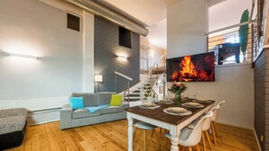A modern living room with a dining area, wooden flooring, a grey sofa with a green pillow, a TV mounted on the wall, and a staircase leading to an upper level. - Spacious ground floor loft with 140 m² - ideal for families & groups of up to 6 (7) people (Magdeburg)