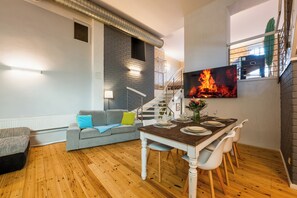 Un salon moderne avec un coin repas, un plancher en bois, un canapé gris agrémenté d’un coussin vert, un téléviseur fixé au mur et un escalier menant à un niveau supérieur.