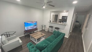 Una sala de estar moderna con un sofá verde, una mesa de centro de madera, un televisor sobre un mueble bajo, un ventilador de techo y una zona de cocina con taburetes de bar.