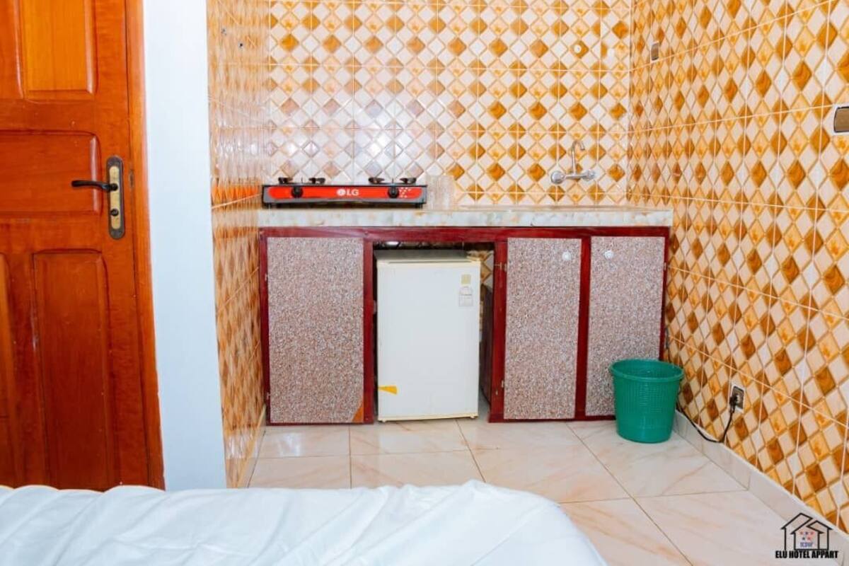 Quarto pequeno com uma cama, cozinha compacta com fogão a gás, geladeira e lixeira.