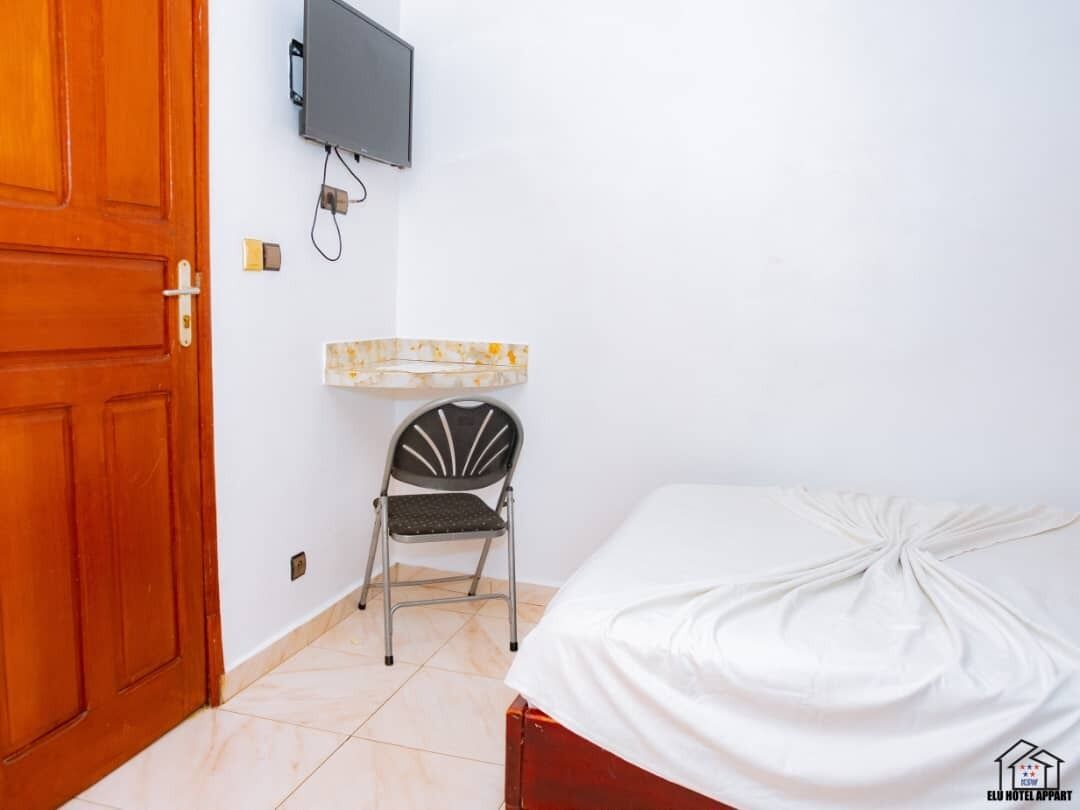 A small room with a wooden door, a bed with a white sheet, a small table with a marble-like surface, a chair, and a mounted television.