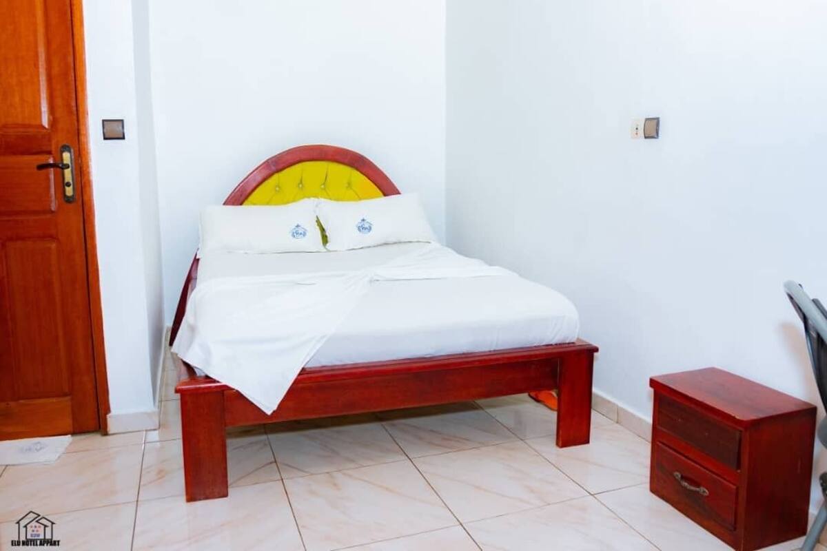 Uma cama de solteiro com estrutura de madeira e cabeceira amarela, um colchão branco e duas almofadas com logotipo. Um criado-mudo de madeira com uma gaveta está à direita da cama.
