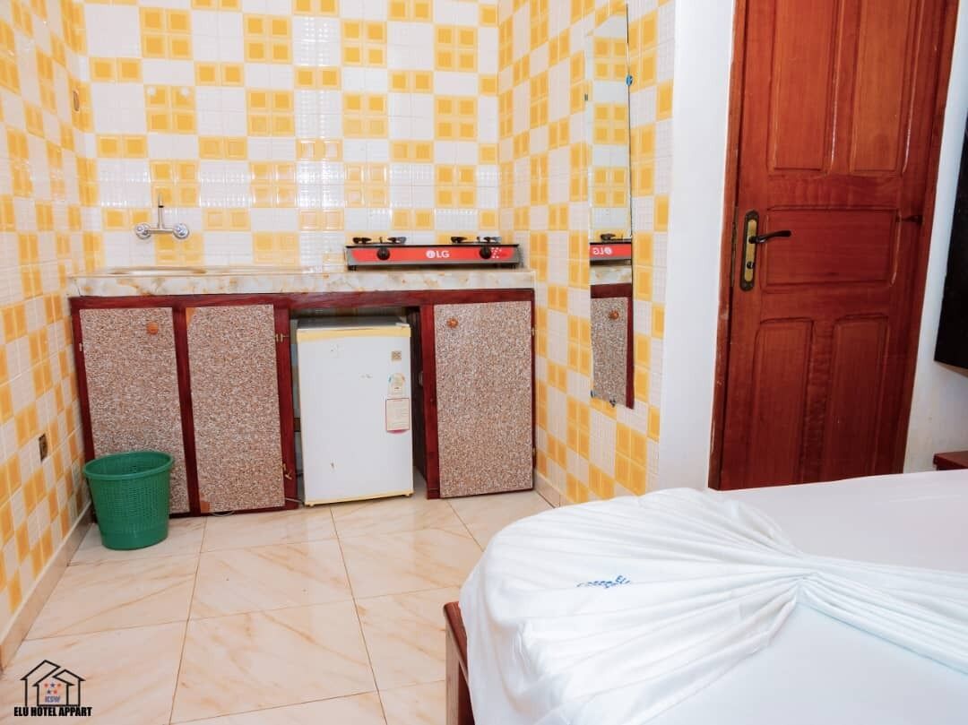 Une petite chambre d’hôtel avec un lit, une kitchenette équipée d’une cuisinière LG, une poubelle verte et une porte en bois.