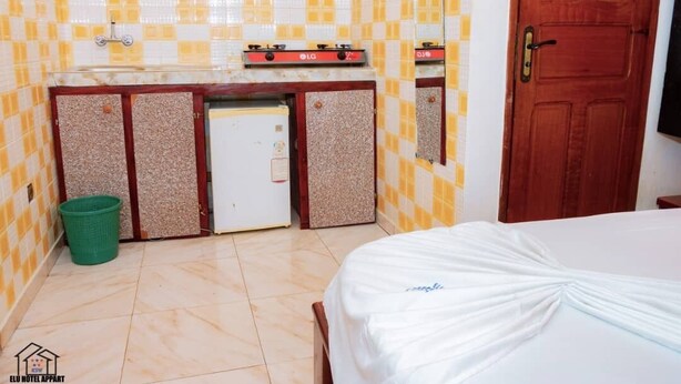 A small hotel room with a bed, a kitchenette with an LG stove, a green trash bin, and a wooden door. - Elu Hotel Appart (Douala)
