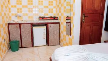 Une petite chambre d’hôtel avec un lit, une kitchenette équipée d’une cuisinière LG, une poubelle verte et une porte en bois.