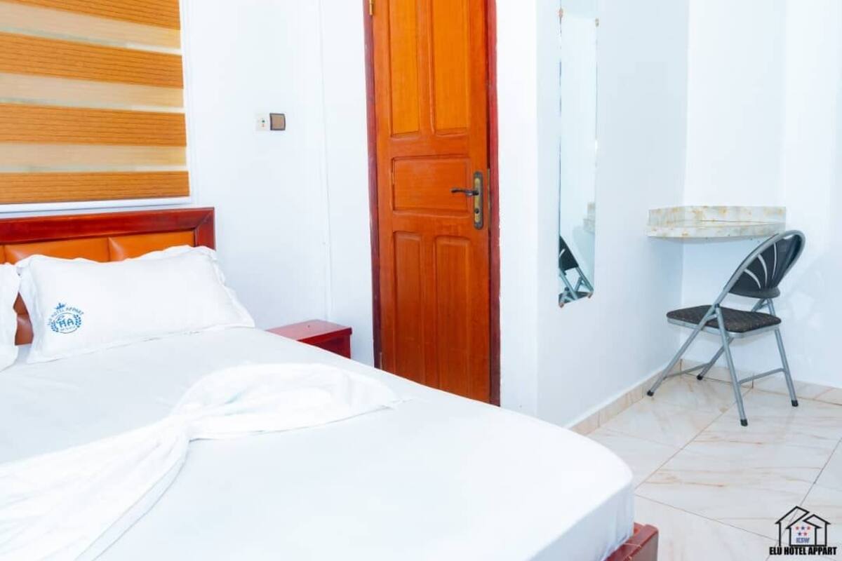 Quarto de hotel com cama, porta de madeira, mesa pequena, cadeira e espelho na parede.