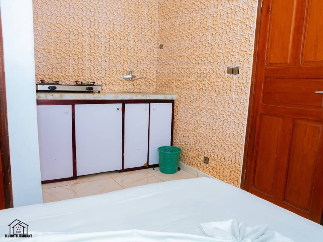 A small, compact room with a kitchenette, a green trash bin, and a wooden door.