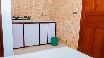 A small, compact room with a kitchenette, a green trash bin, and a wooden door.