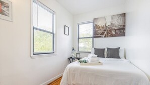 Une chambre à coucher avec un lit, deux fenêtres, une table de chevet et un tableau représentant un paysage urbain.