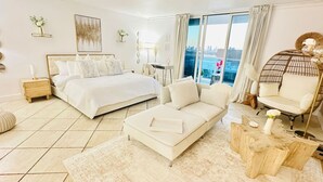 Une chambre moderne avec un grand lit, un canapé, un fauteuil et une vue sur la ville grâce à de grandes fenêtres.
