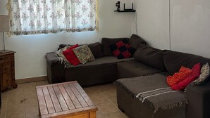 A living room with a sectional sofa, a wooden coffee table, a window with floral curtains, and a wall-mounted shelf. - Cantinho das Emerências, a house near the beach in a rustic style with a swimming pool! (Armação dos Búzios)