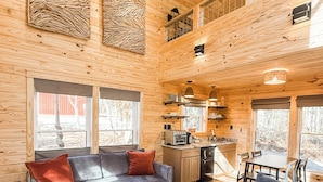 Ruim interieur van een chalet met een hoge plafond, een gezellige zithoek met een bank en fauteuils, een keuken met een eettafel, en grote ramen met uitzicht op de omgeving.