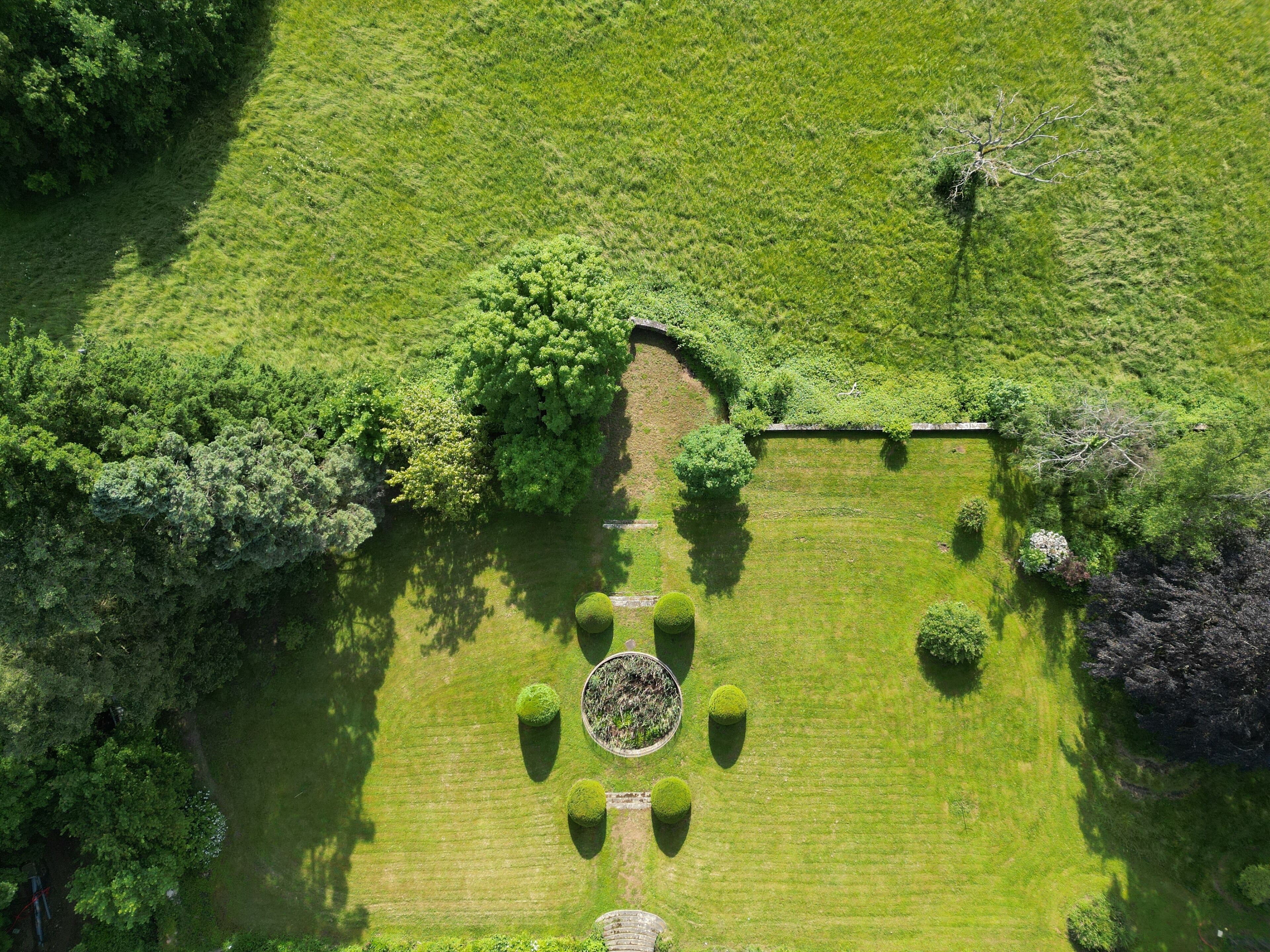 An aerial view of a well-maintained garden with a central circular lawn, surrounded by dense greenery and a stone wall.