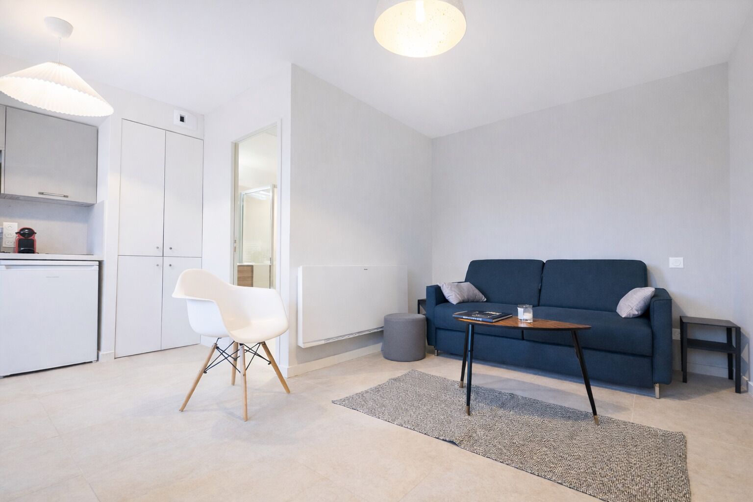 A modern living room with a blue sofa, a white chair, a small table, and a kitchen area with cabinets and a refrigerator.