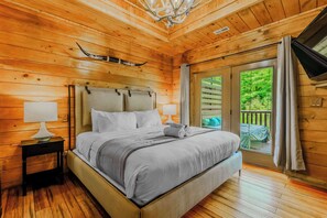 A wooden cabin bedroom with a bed, nightstand, lamp, and a view of a deck. - Dollywood & Pigeon Forge | Pool access | Sleeps 12 (Pigeon Forge)