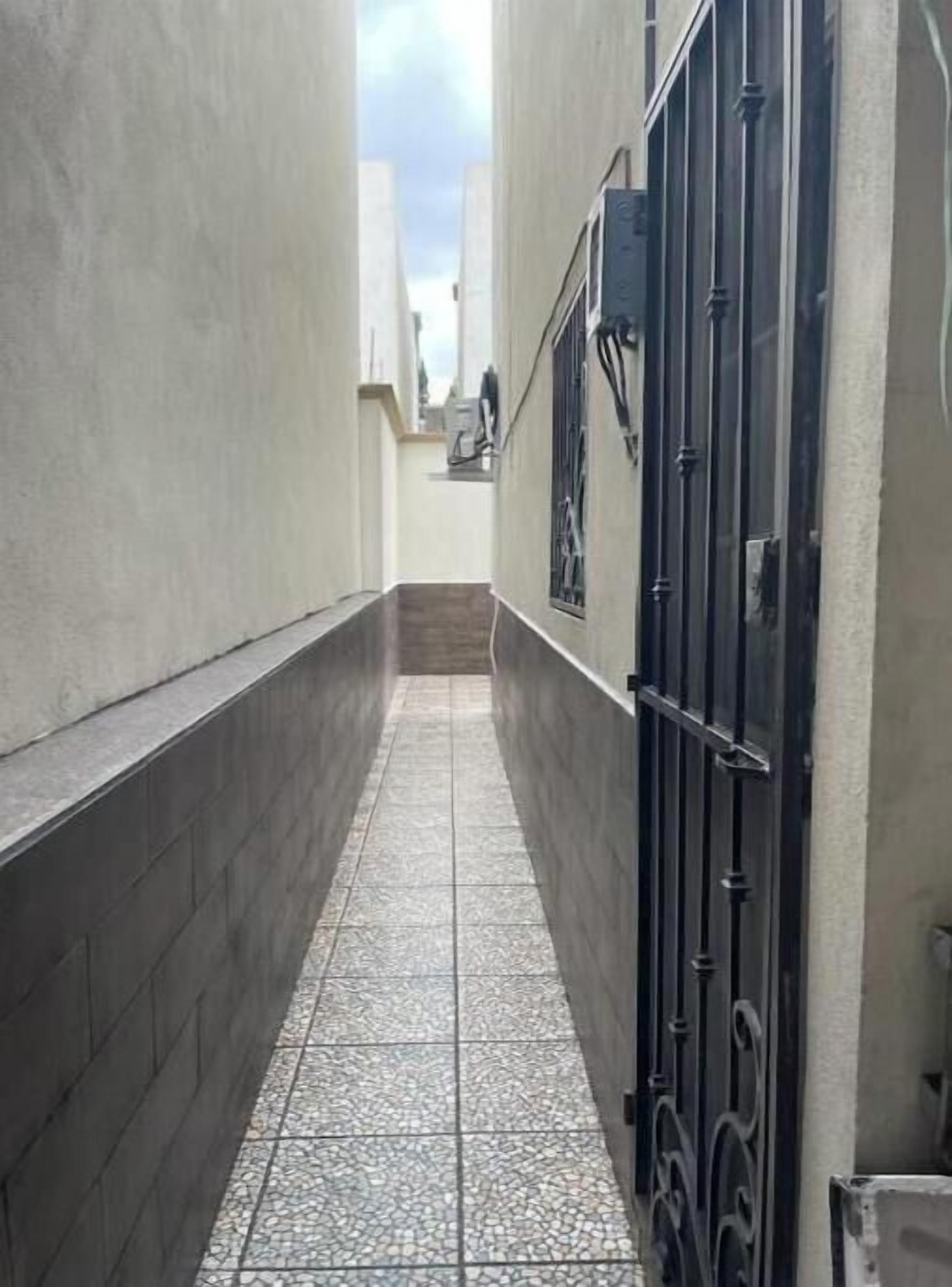 Un estrecho pasillo con piso de baldosas, un portón negro y una persona a lo lejos.