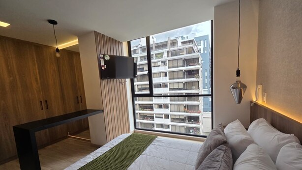 Ein modernes Schlafzimmer mit einem großen Bett, einem Holzschrank, einem an der Wand montierten Fernseher und Blick auf ein Hochhaus.