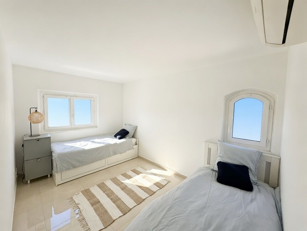 A small, bright bedroom with two beds, a bedside table, a lamp, and a window.