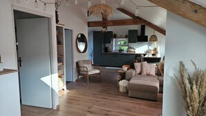 A cozy living room with a sofa, a chair, and a dining area with a table and chairs. There is a kitchen in the background with cabinets and a window. - Charming cocoon in the heart of the spa district (Vittel)